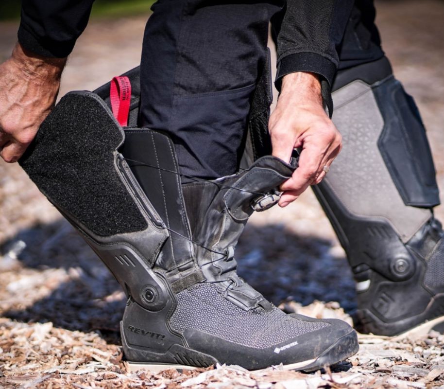 Close-up of a rider adjusting REV’IT! Expedition GTX boots with BOA dial and Velcro flap, showing armored shin and ankle protection.