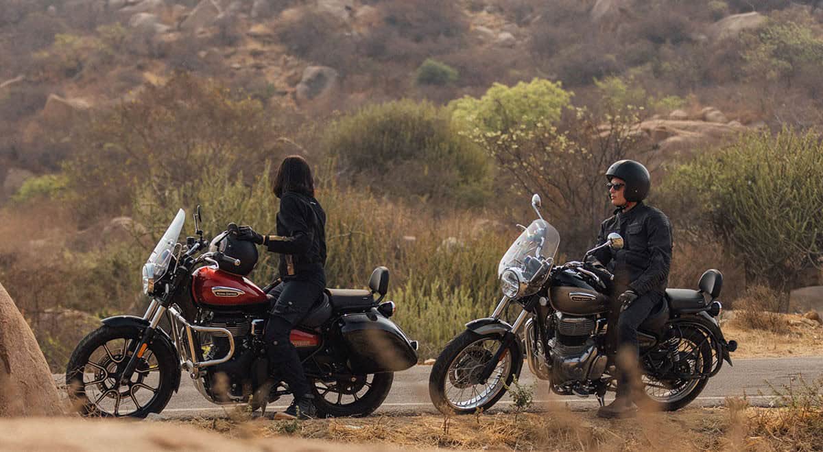 Two riders on Royal Enfield Meteor 350 motorcycles paused on a scenic country road surrounded by hills