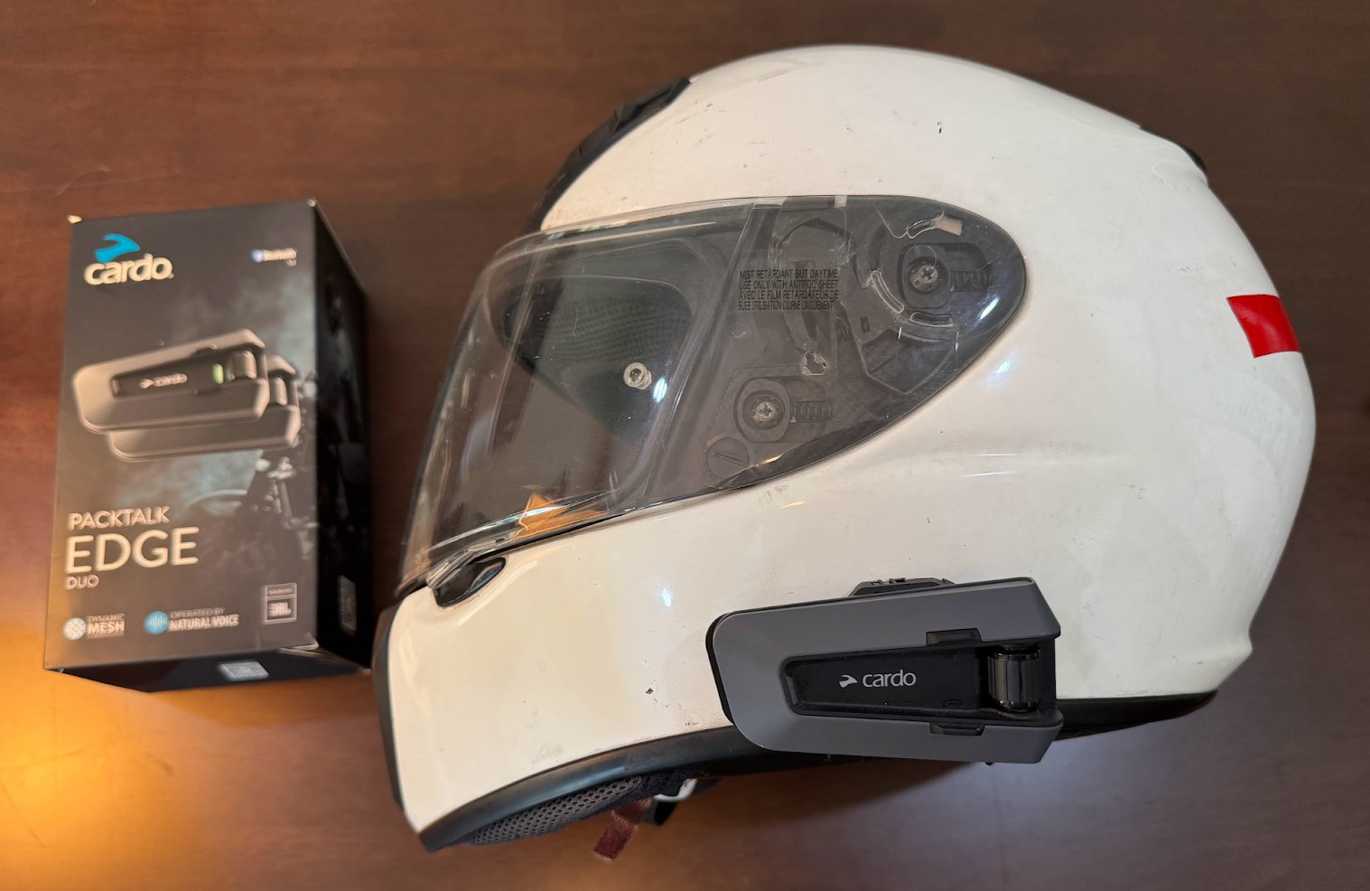 White motorcycle helmet fitted with a Cardo Packtalk Edge communicator beside its retail box, showing mesh and JBL features.