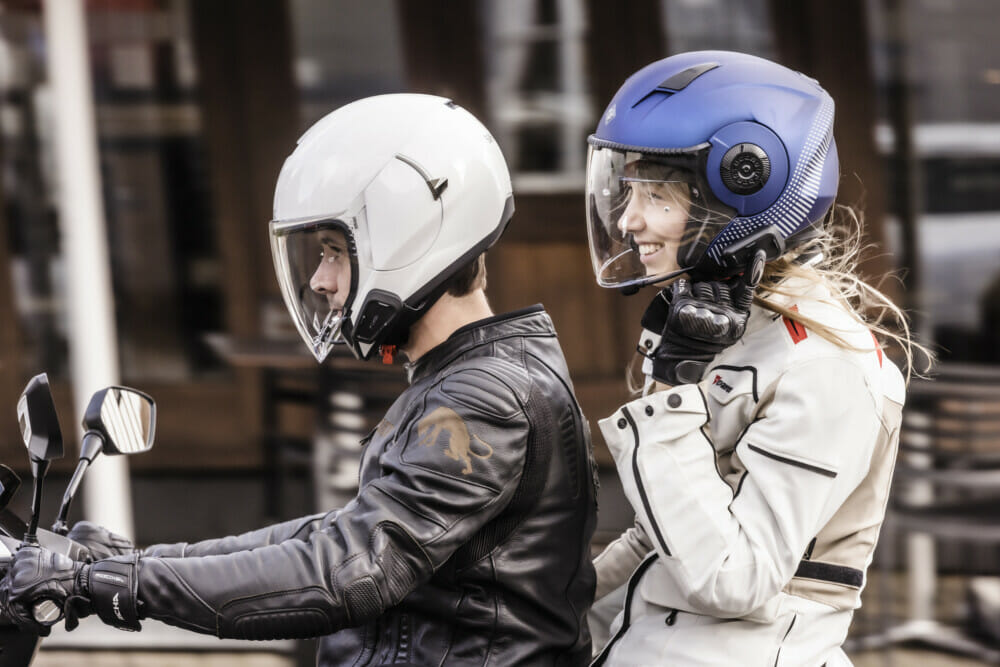 Two riders wearing helmets with Cardo Spirit communicators, smiling while riding together through the city on a scooter.