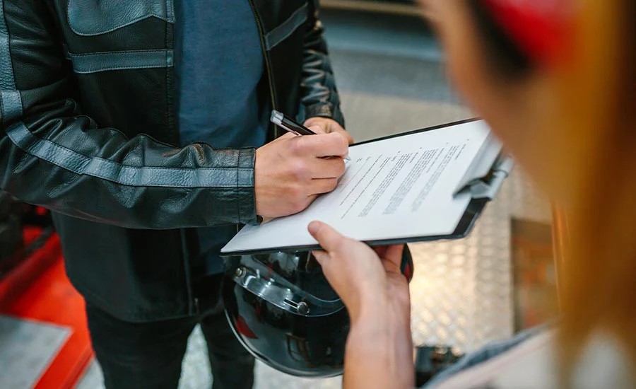 Motorcyclist in a leather jacket signing insurance paperwork while holding a helmet, representing motorcycle insurance costs and coverage.