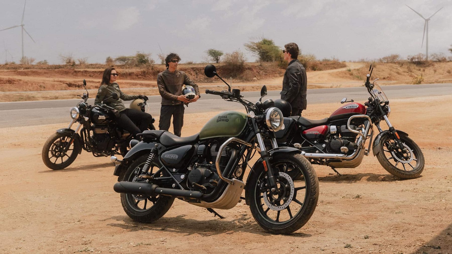 Group of riders chatting beside their Royal Enfield cruisers on a sunny open road with wind turbines in the background