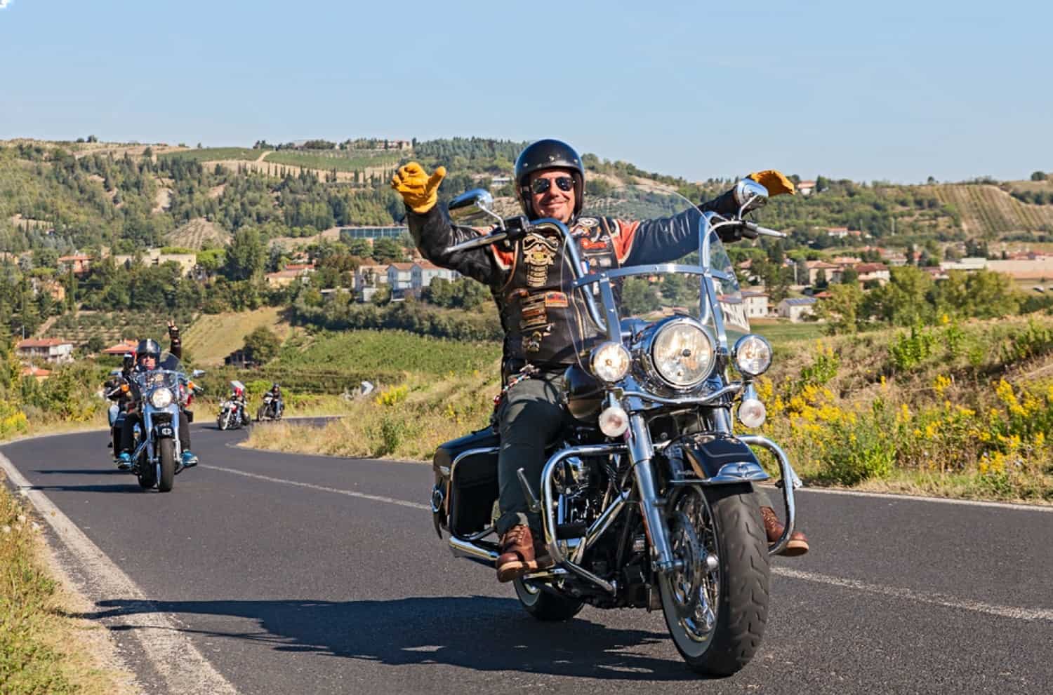 Group of motorcycle riders cruising through scenic countryside roads on a sunny day, smiling and waving in full touring gear.