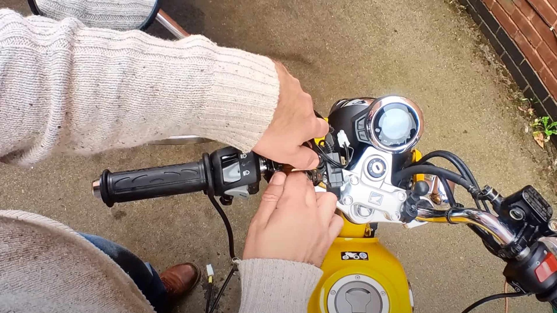 Close-up of a rider installing motorcycle grips and wiring on a yellow bike, showing the handlebar controls and wiring setup.