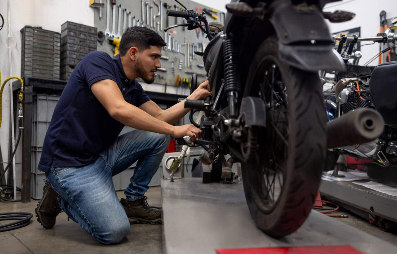 Mechanic installing or adjusting a motorcycle tire in a workshop with tools on the wall in the background.