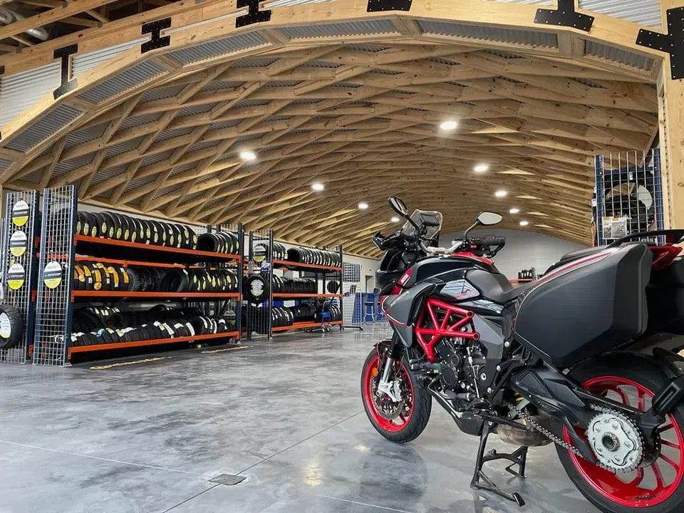 Motorcycle inside a dealership workshop with racks of tires and modern wooden roof architecture in the background.
