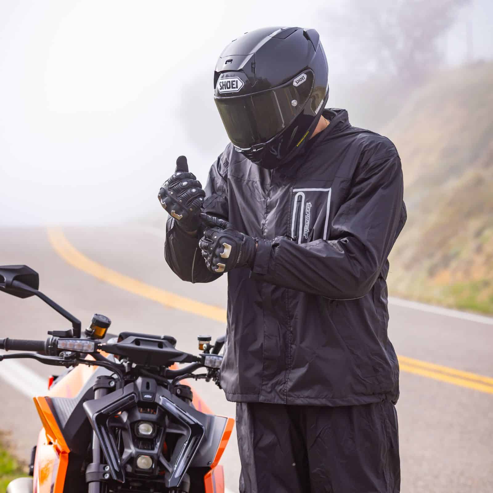 Rider wearing the Tourmaster Defender two-piece rainsuit adjusting gloves beside a motorcycle on a foggy road