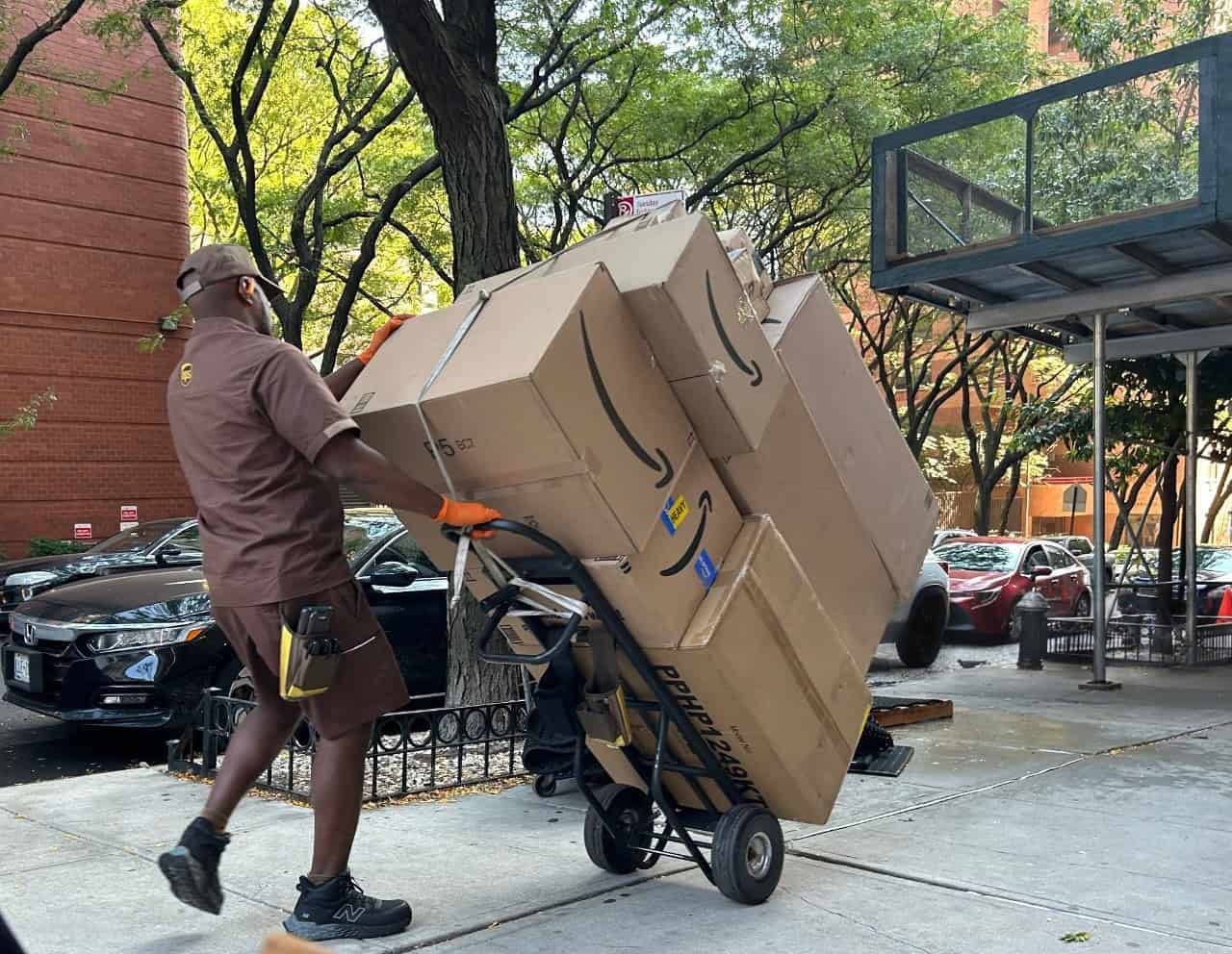 UPS driver transporting large Amazon boxes on a hand truck along a city sidewalk during a delivery.