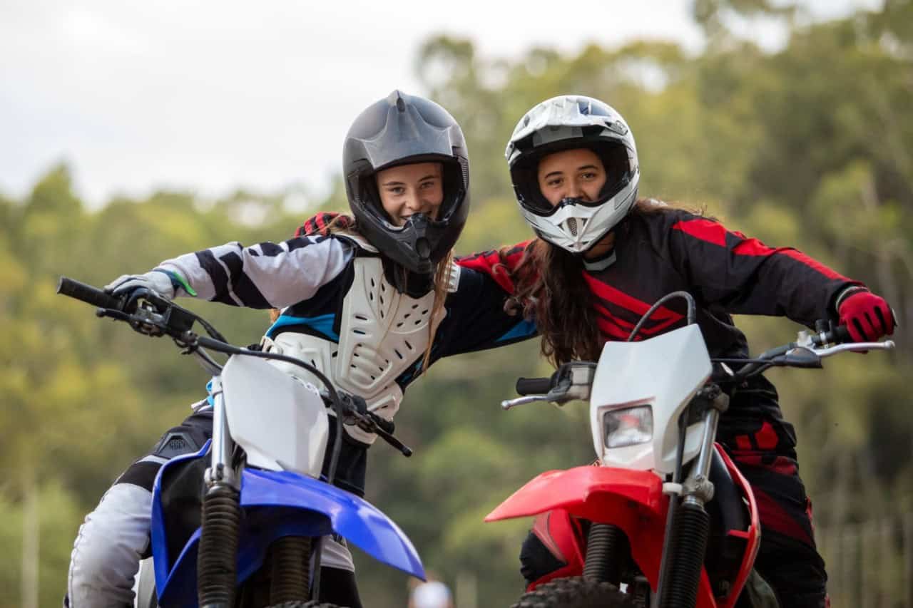 Two young motorcyclists in helmets and riding gear smiling together on their bikes, symbolizing how age and experience affect insurance costs.