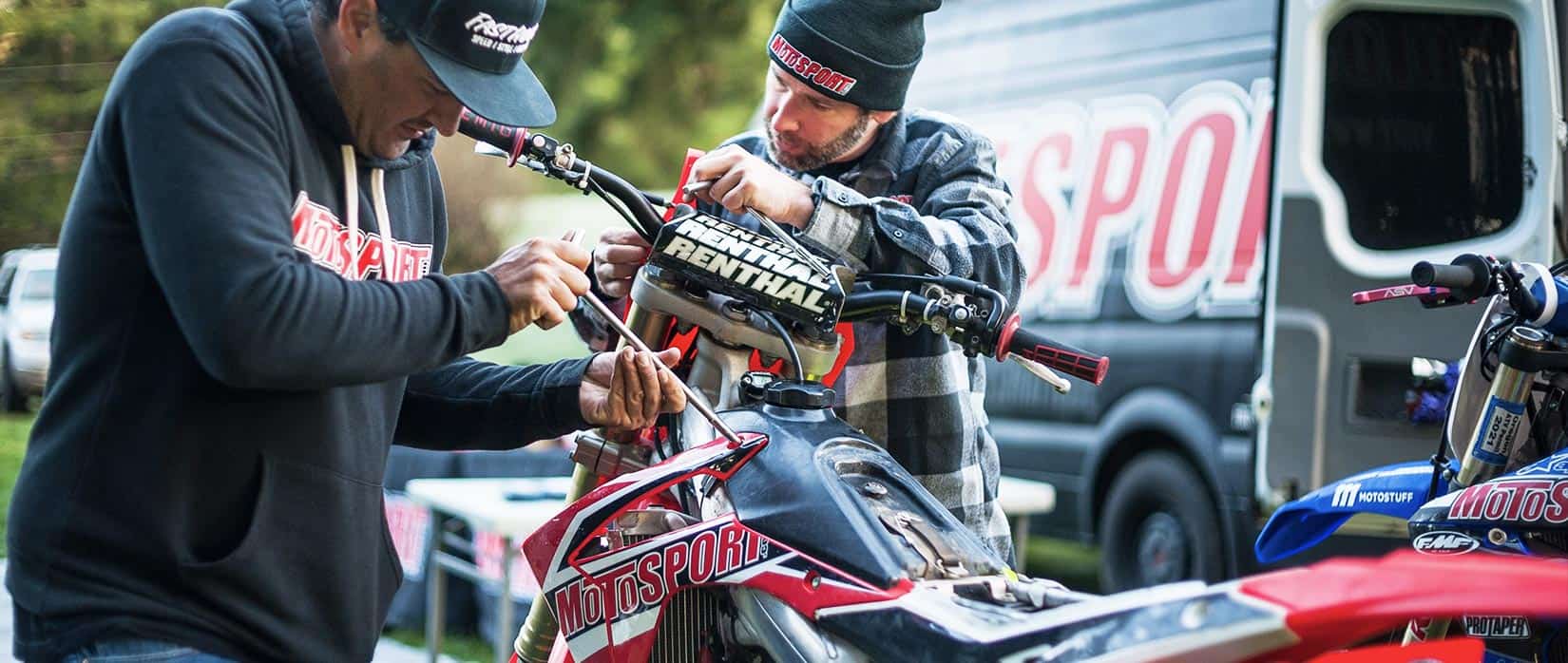 Two MotoSport team members working on dirt bikes outdoors, wearing MotoSport gear with a branded van in the background.