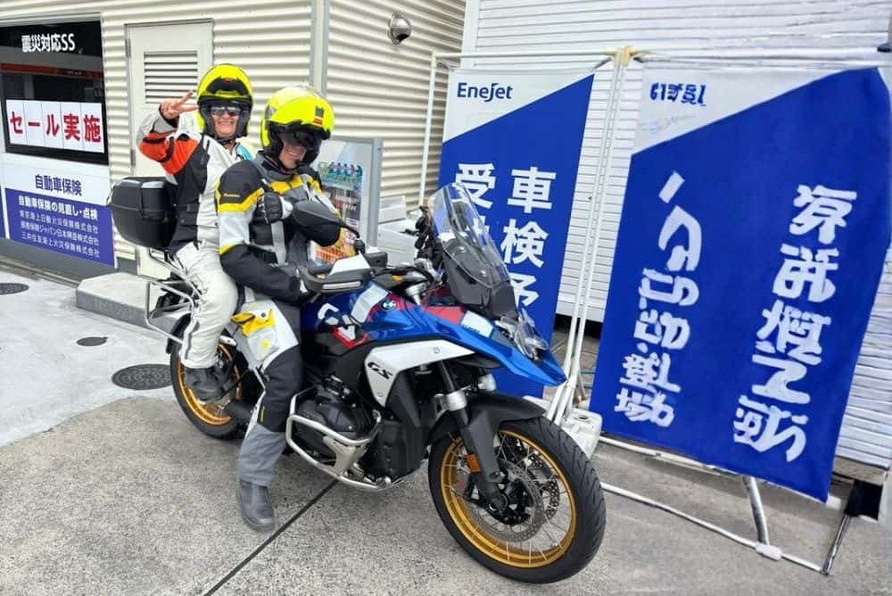 Two riders in full gear on a BMW adventure motorcycle at a gas station, smiling and ready for the road