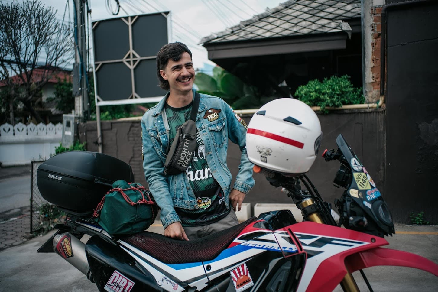 Smiling motorcyclist standing beside his packed adventure bike with a helmet resting on the handlebar.