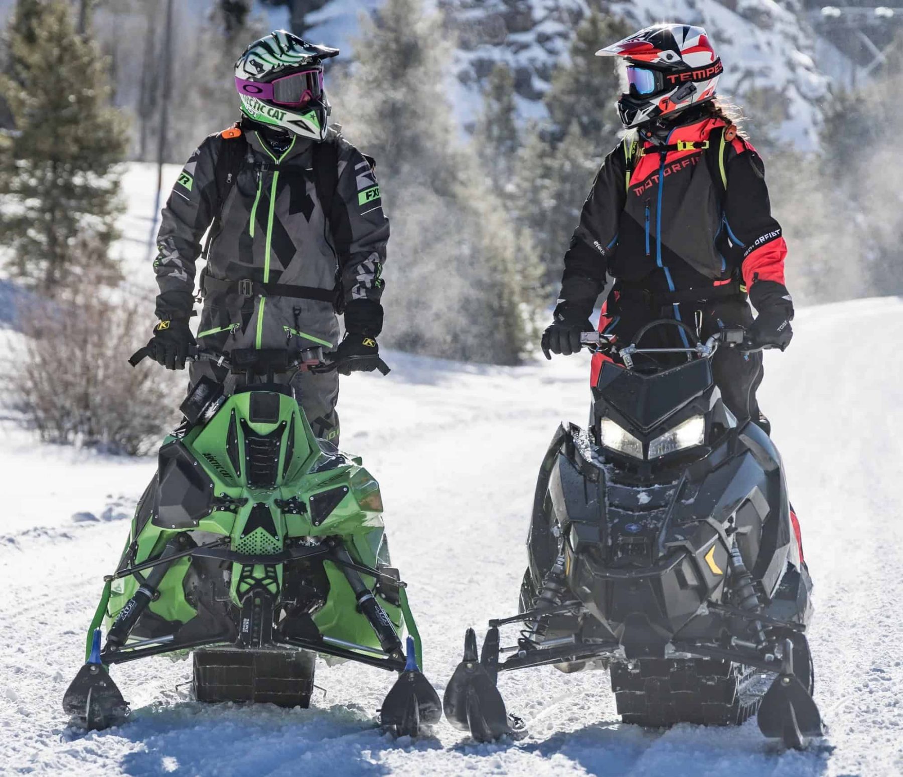 Two snowmobile riders wearing helmets and gear, preparing to ride together on a snowy trail