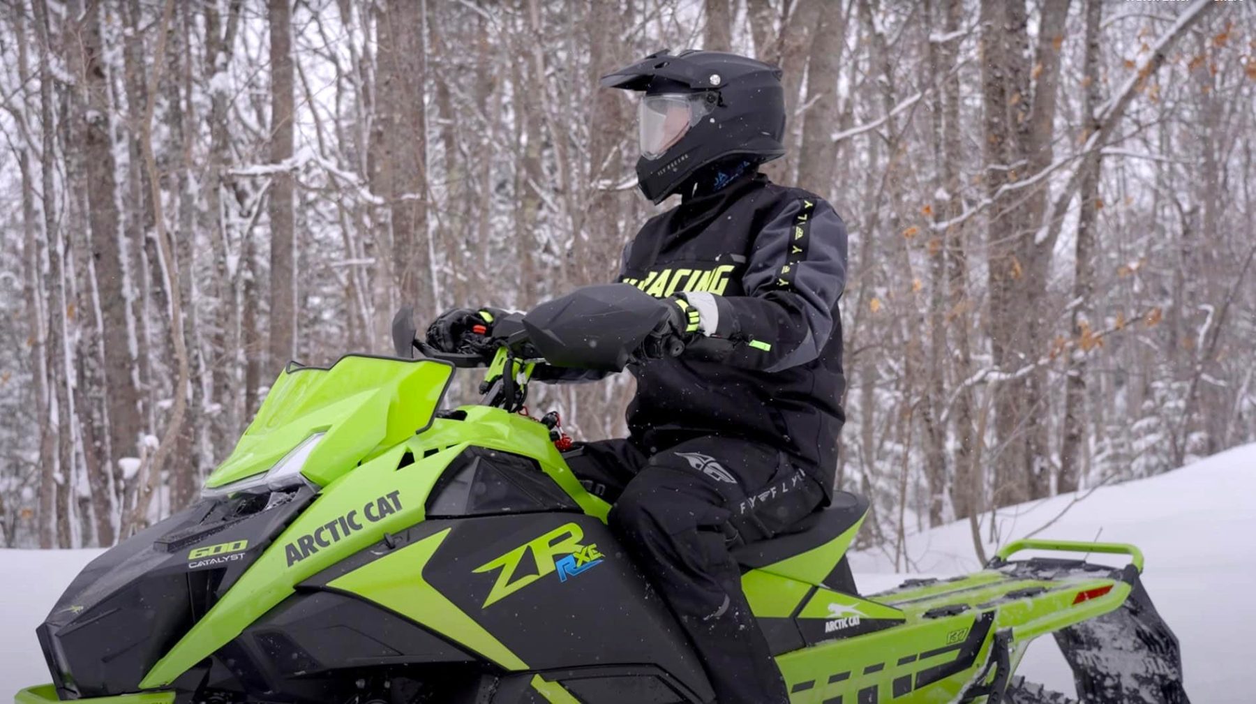 Rider on a bright green Arctic Cat snowmobile wearing winter riding gear in snowy woods