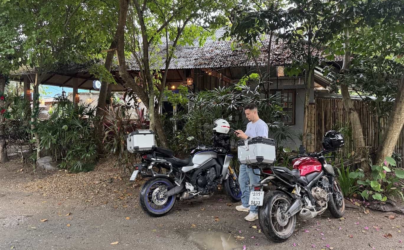 Man checking his phone beside two adventure motorcycles parked outside a rustic roadside café surrounded by lush greenery.