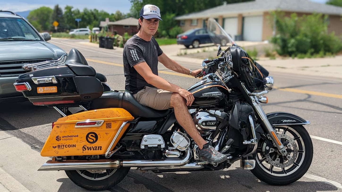 Man sitting on a Harley-Davidson touring motorcycle on a sunny street, wearing a casual outfit and cap.