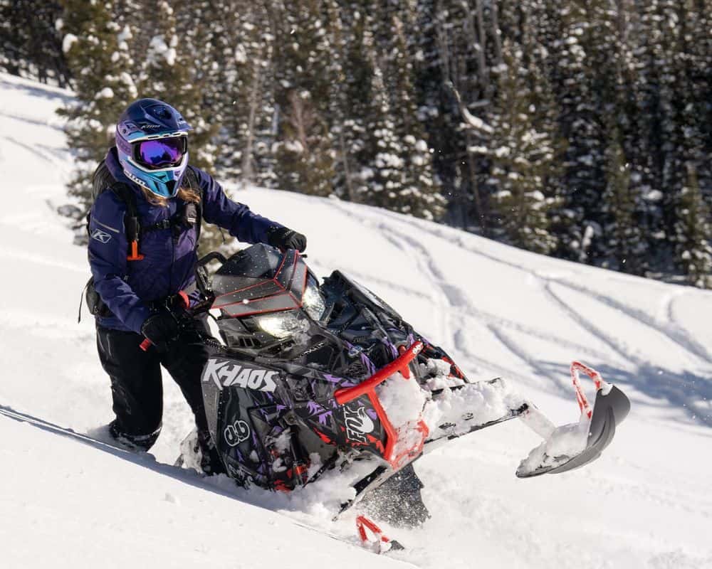 Woman riding a snowmobile uphill wearing the Klim Alpine Women’s Jacket in deep mountain snow