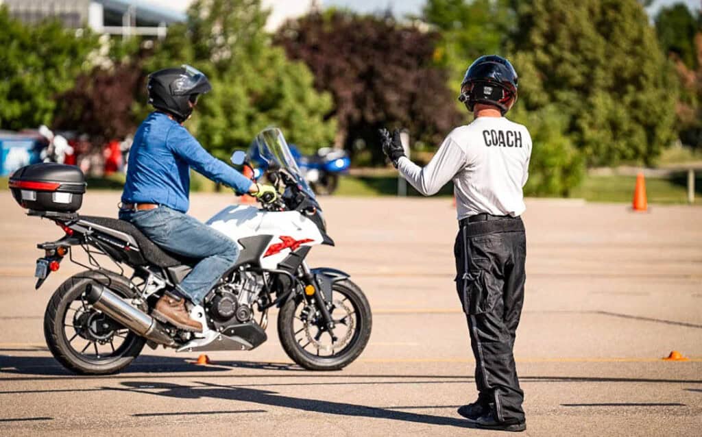 Motorcycle safety coach training a new rider in a parking lot course, illustrating discounts for instructors and club members.