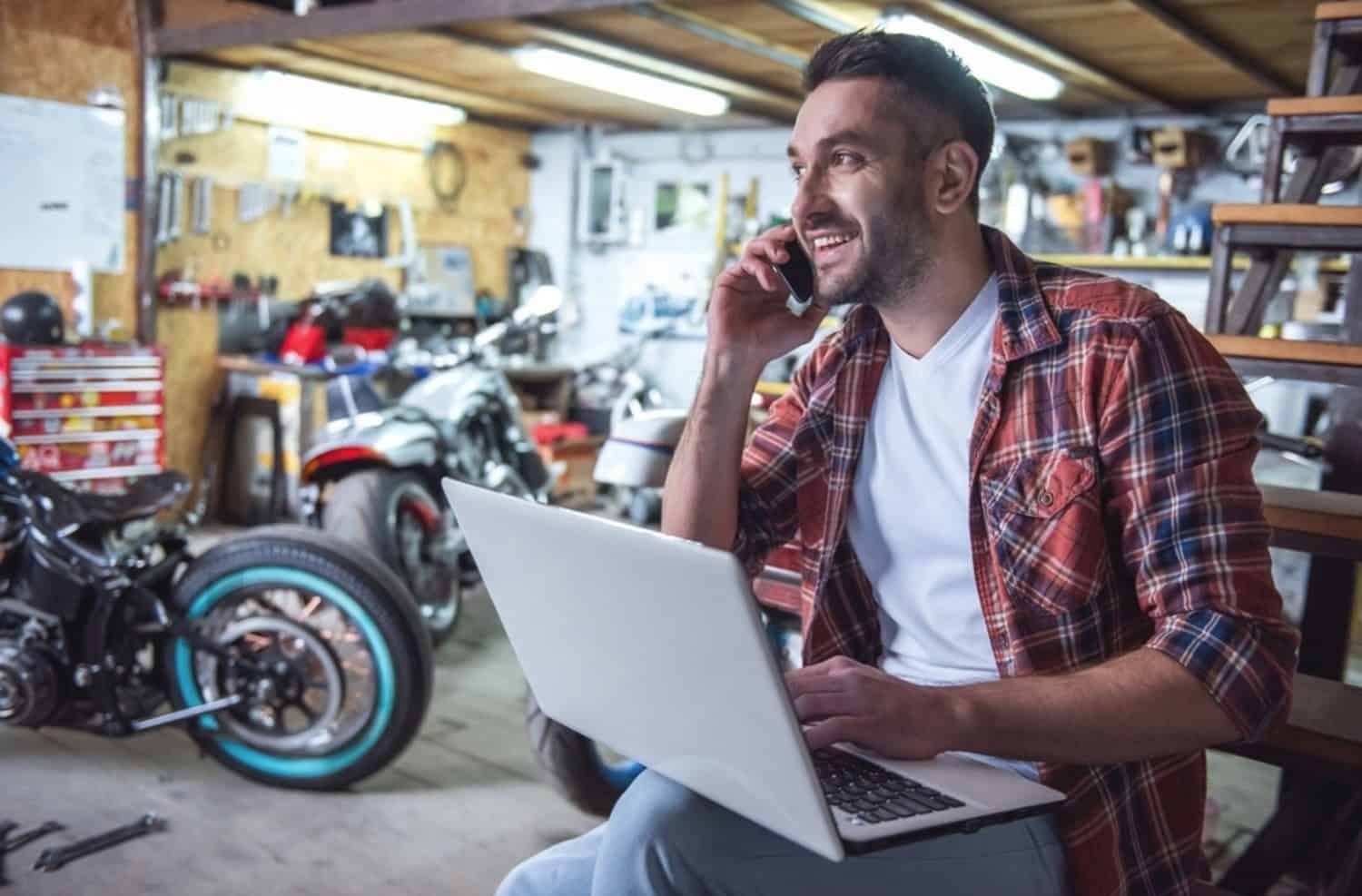 Smiling motorcyclist in a garage using a laptop and phone to compare motorcycle insurance quotes online.