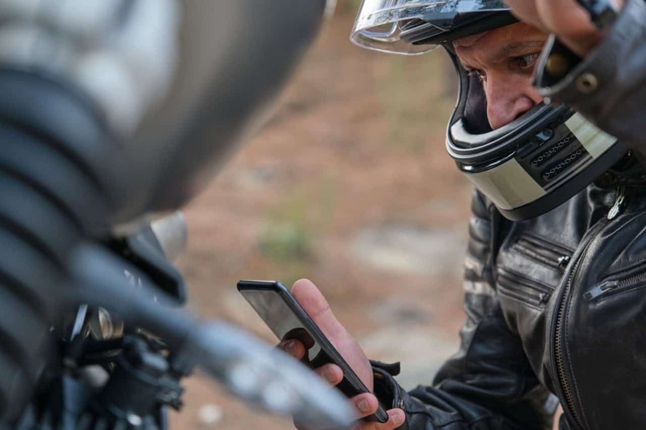 Close-up of a motorcyclist in a helmet and leather jacket checking his phone beside his bike.