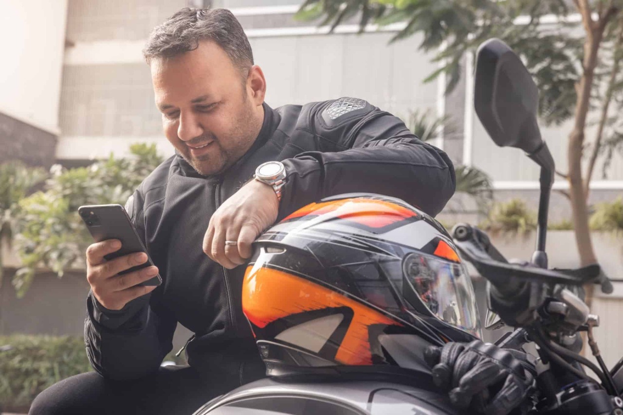 Motorcyclist in black riding gear smiling while checking his phone beside his helmet and bike.