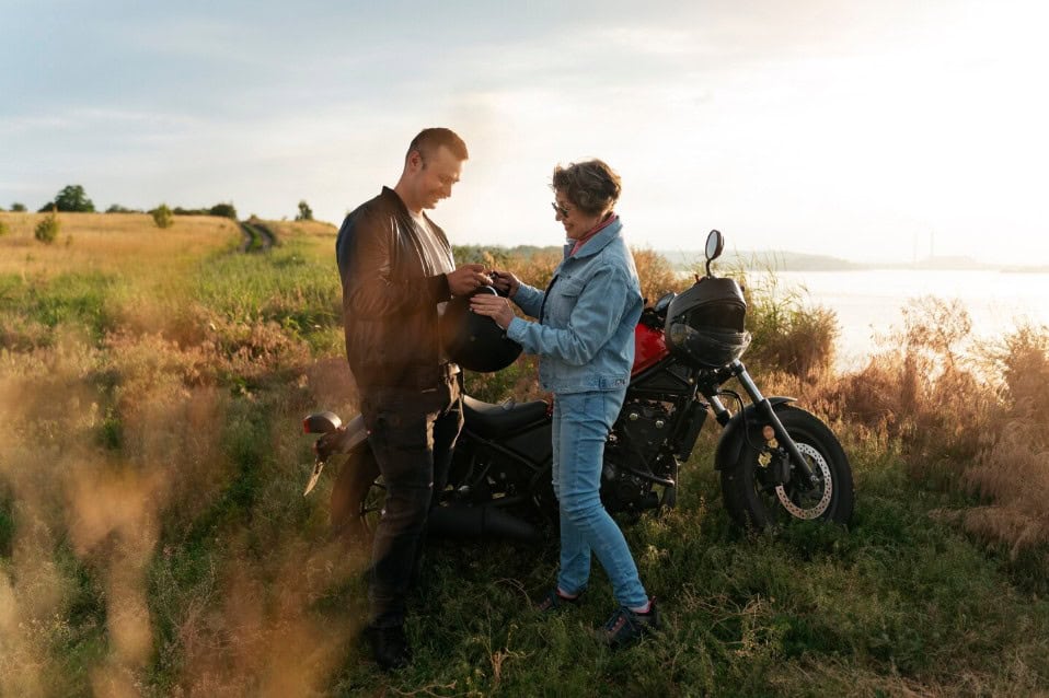 Two motorcyclists sharing a helmet beside a parked bike in a sunny field