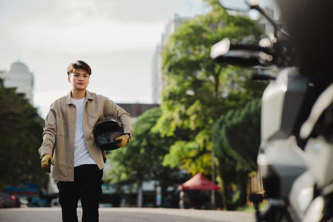 Young man holding a motorcycle helmet and walking toward his bike, symbolizing new riders learning that clean records don’t always mean low premiums.