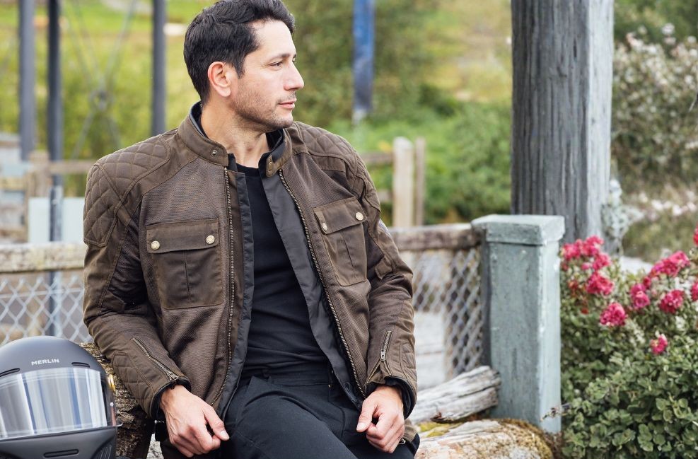 Man wearing heritage-style motorcycle jacket sitting outdoors with helmet beside him