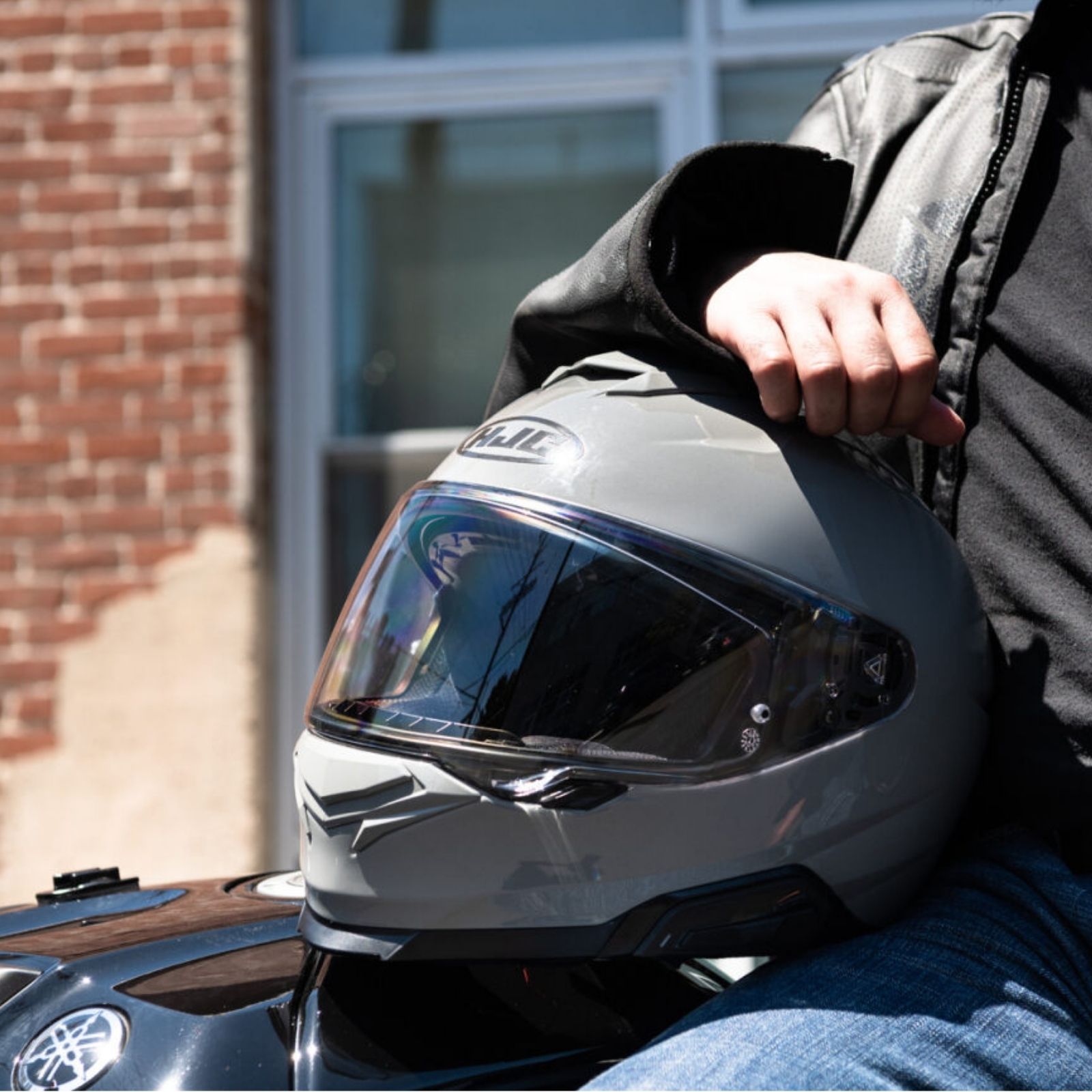 Rider holding a matte gray full-face HJC helmet with tinted visor while sitting on a motorcycle outdoors