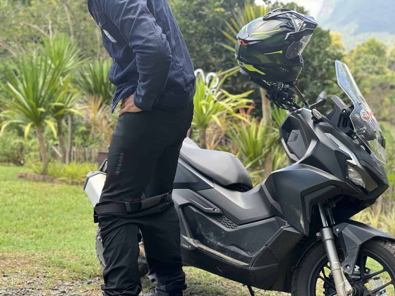 Rider wearing protective base layer pants under outer clothing beside a scooter in a tropical outdoor setting